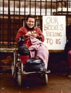 Una bella immagine di Gabriella Bertini che lavora a maglia mentre partecipa ad una manifestazione (dietro di lei un cartello con la scritta, in inglese, “I nostri corpi ci appartengono”).