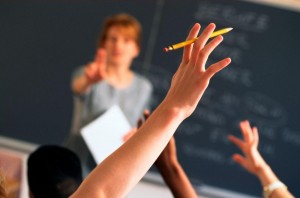 In una classe si intravede un’insegnante fuori fuoco, in primo piano la mano alzata di uno studente con una matita chiede la parola.