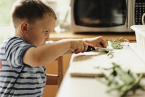 Un bambino, munito di coltello, pulisce dei fagiolini sopra un tagliere (foto di Ksenia Makagonova). 