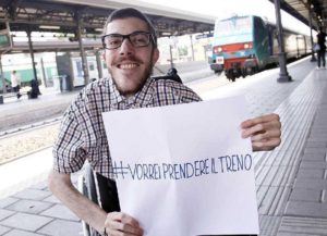 Iacopo Malio sui binari, alla stazione dei treni, mostra un cartello con la scritta “#vorrei prendere il treno”, la campagna di sensibilizzazione per l’abbattimento delle barriere architettoniche lanciata nel 2014.