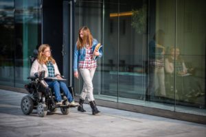 Due giovani studentesse camminano per strada, una di esse ha la sclerosi multipla e si sposta in sedia a rotelle.