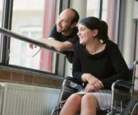 Un uomo ed una donna sorridono mentre guardano fuori da una finestra. Lei è in sedia a rotelle. 