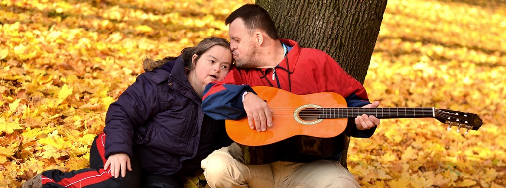 Una donna ed un uomo, entrambi con disabilità intellettiva, in atteggiamento affettuoso, seduti su un mare di foglie gialle, con la schiena poggiata ad un tronco d’albero ed una chitarra in mano.