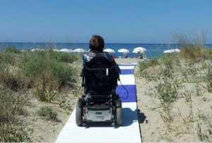 Una persona che si sposta in sedia a rotelle ritratta di spalle mentre percorre una passerella che la conduce in spiaggia. È l’immagine che si trova nell’home page del sito dell’Agenzia per la Vita Indipendente di Roma.