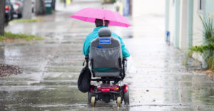 Una persona con disabilità motoria ritratta di spalle mentre, con la sua sedia a rotelle, si sposta sotto la pioggia.