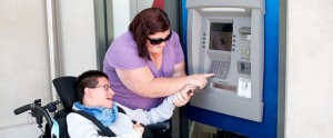 Una persona con disabilità motoria utilizza un bancomat con il supporto della sua assistente personale.