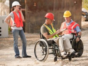 Due lavoratori con disabilità motoria ed una lavoratrice in un cantiere, con i caschi e i giubbotti protettivi. 