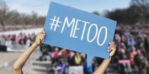 Un cartello con la scritta #METOO è sorretto da due braccia femminili. Sullo sfondo, fuori fuoco, una moltitudine di persone intervenute ad una manifestazione contro la violenza sulle donne.