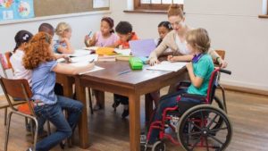 Un gruppo classe composto da diversi/e studenti, di cui una con disabilità motoria, svolge un compito con la supervisione dell’insegnante.
