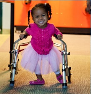 Una bambina con disabilità motoria sorride mentre si sposta con l’ausilio di un deambulatore. 