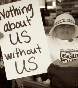 Foto in bianco nero di una donna con disabilità che manifesta per i propri diritti esibendo un cartello con la scritta “Nothing about us without us” (Nulla su di noi senza di noi).